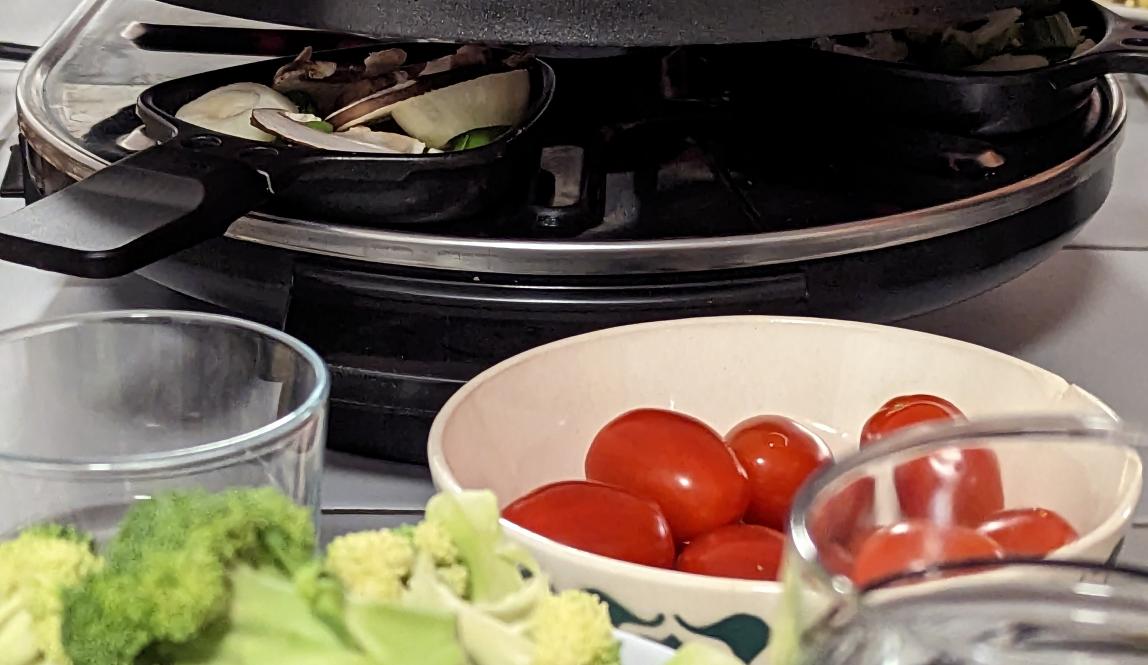 Table with bowl of tomatoes and broccoli in front of a raclette grill. 