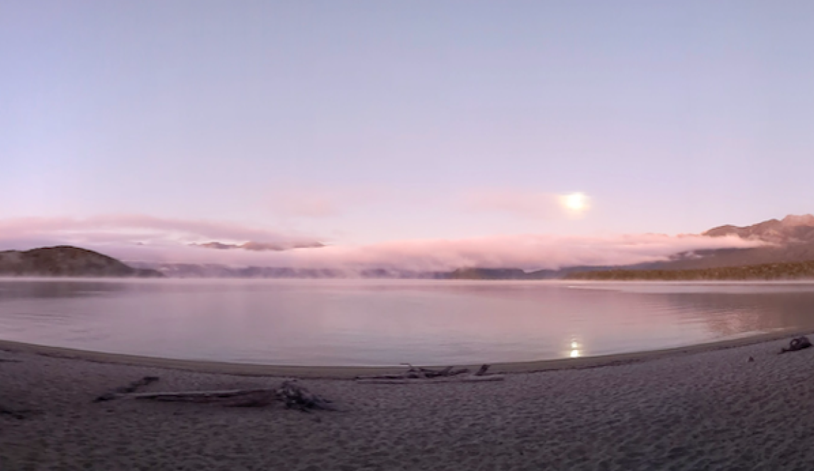 A panoramic view of a lake at dawn with the moon rising over it and mist rising off the water 