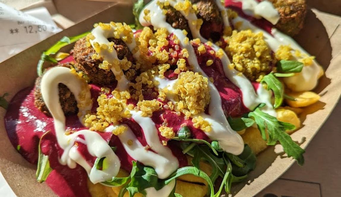 Cardboard plate with poutine with small balls of falafel scattered in it, covered in pink humus and yogurt dressign with arugula