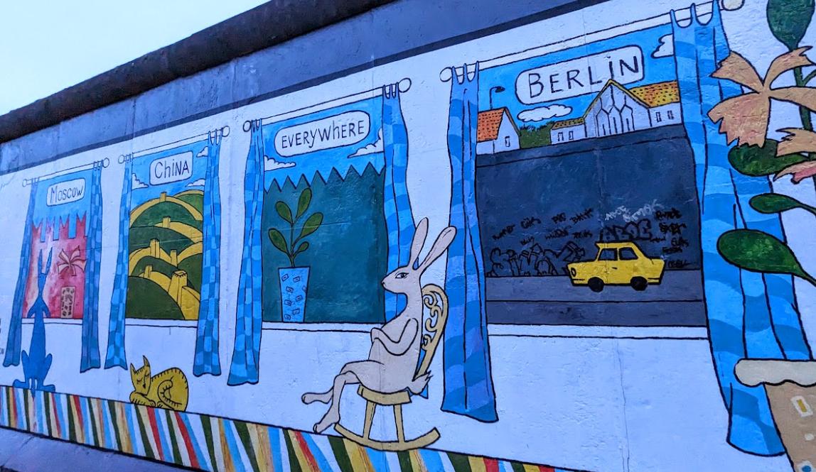 Painting on Berlin Wall stating Berlin with doodle of Berlin wall and taxi Trabi, Everywhere with a plant and wall as well as a bunny, China showing the Great Wall of China with a cat sleeping under it, and Moscow showing the Kremlin and a dog looking at it