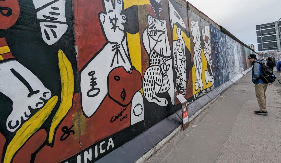 Painting called La Berlinica on Berlin Wall, Wall is stretching to the left and the painting is a red, yellow, and black version of Picasso's La Guernica
