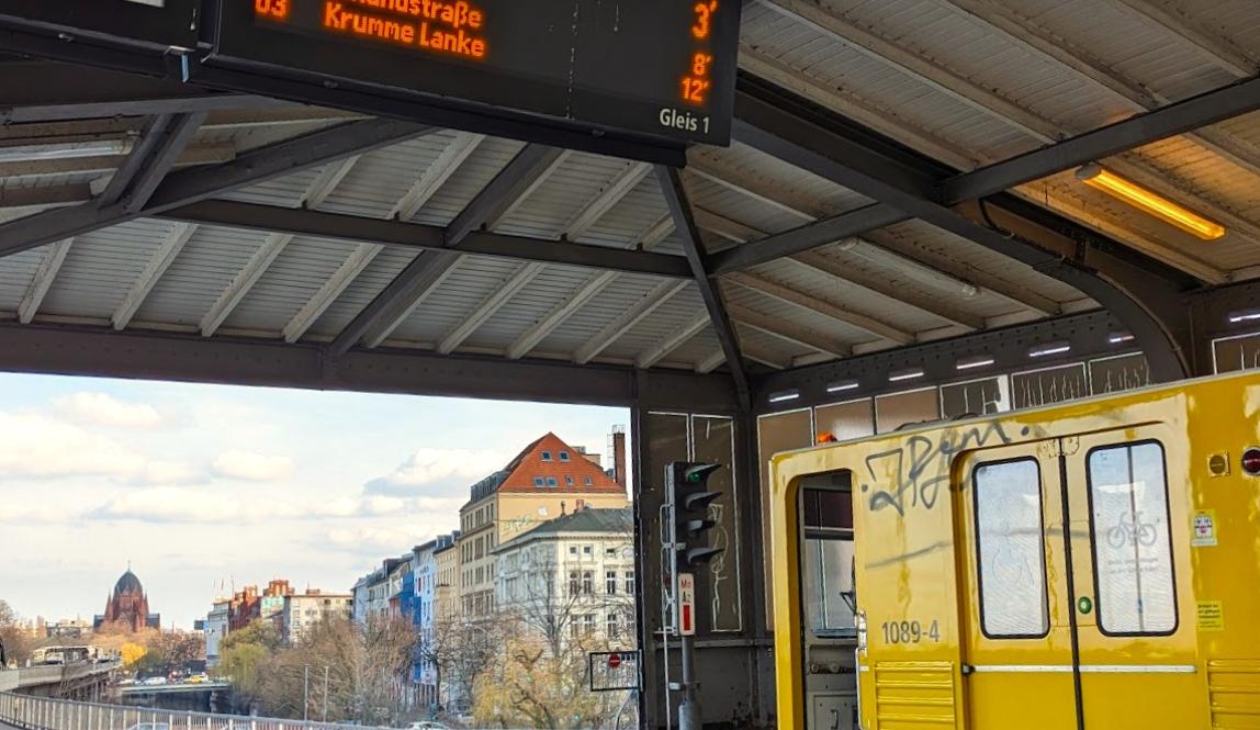 Subway station with yellow subway car with door open on the bottom right, top left corner shows sign with U3 Krumme Lanke and U1 Uhlandstrasse on it, in the background there's the Berlin skyline at twilight