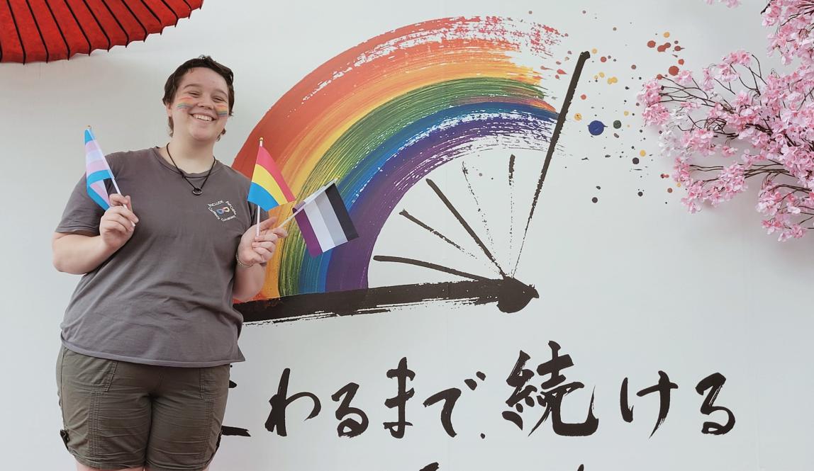 Author, Macks, standing in front of a poster from Tokyo Pride with mini pride flags.