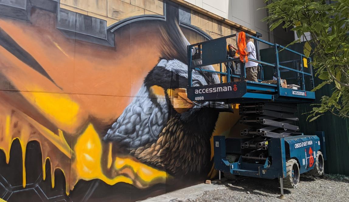 Someone on a construction lift paints a mural of a large orange and grey bird