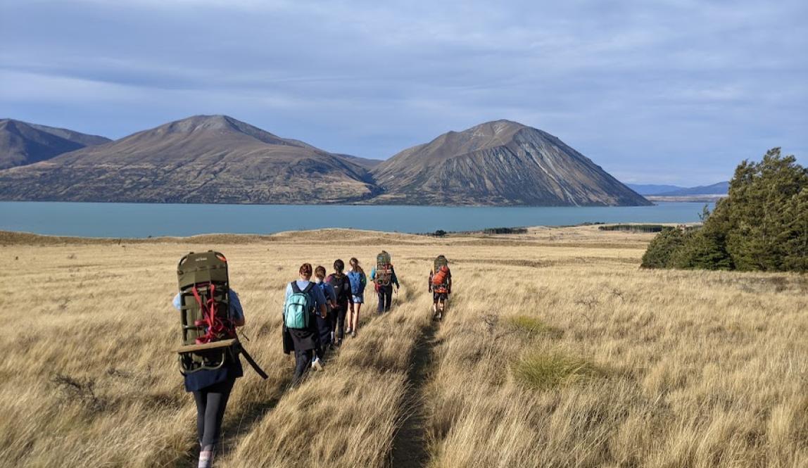 A group of people with backpacks walk down a trail across a plain, with a lake and mountains in the background.