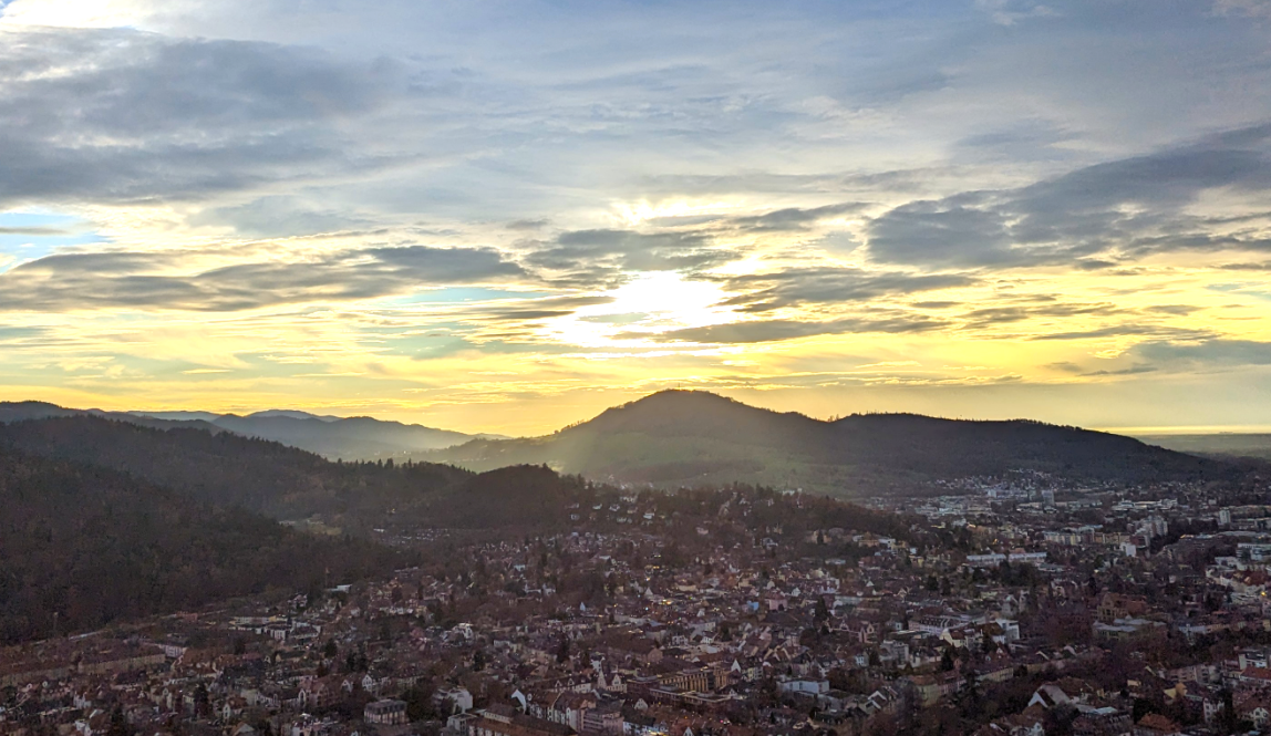 Freiburg at sunset, the sky is a mix of pale yellow and gray-blue due to the clouds, there are black mountains and little houses with some of the glow from the sun on them