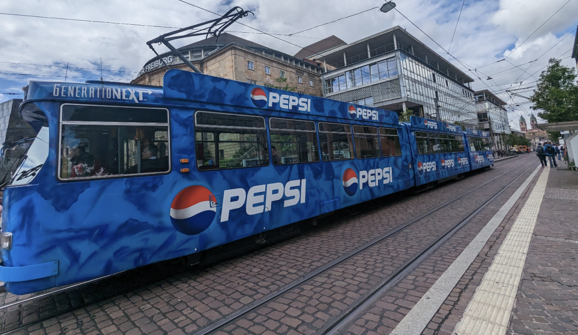 Blue tram with pepsi logo on it heading to the left bottom corner of the image, on rail surrounded by cobblestone road and sky above it is sunny with some clouds