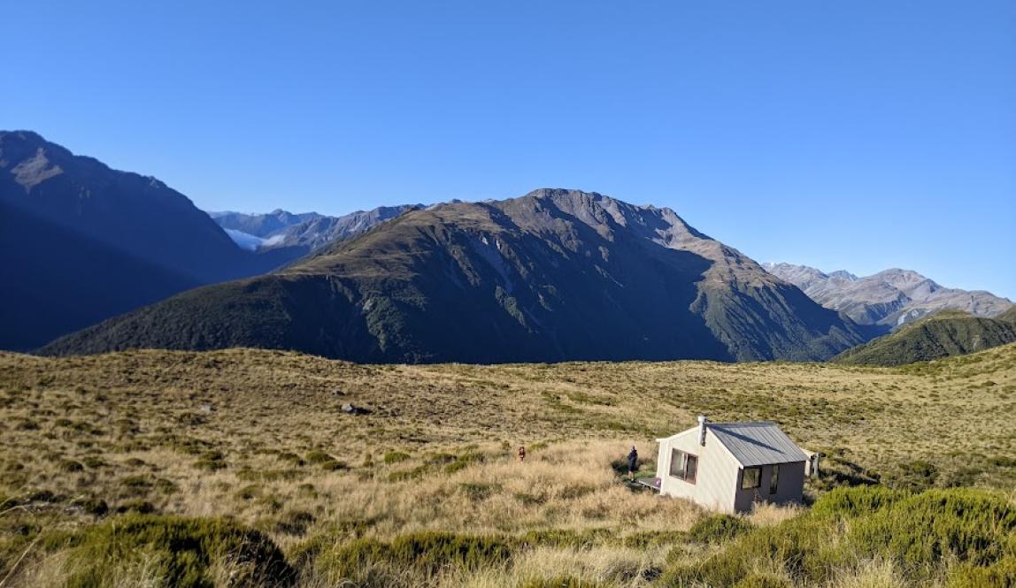 A small hut in a field of tussock on a mountain with the peaks of other mountains in the distance 
