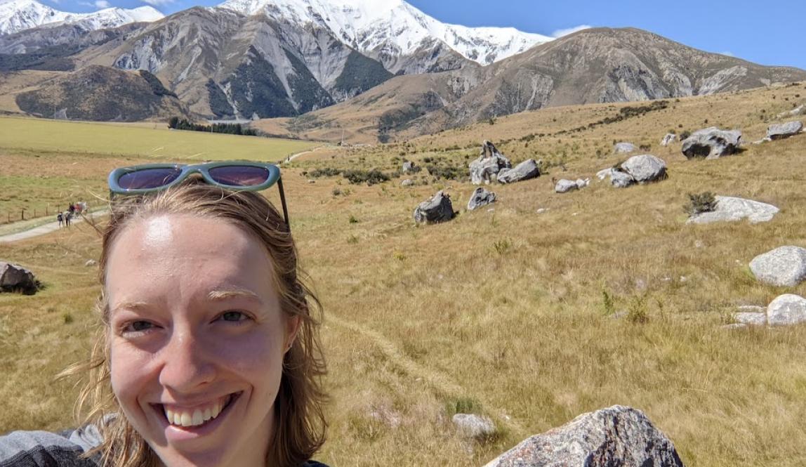 A selfie of me smiling in front of a big field with boulders and snowy mountains in the background