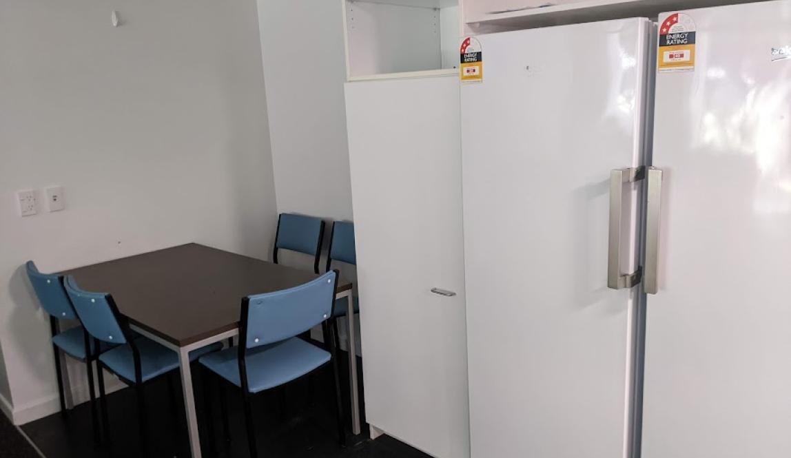A small dining table surrounded by blue chairs, next to a large fridge and freezer and some cupboards. 