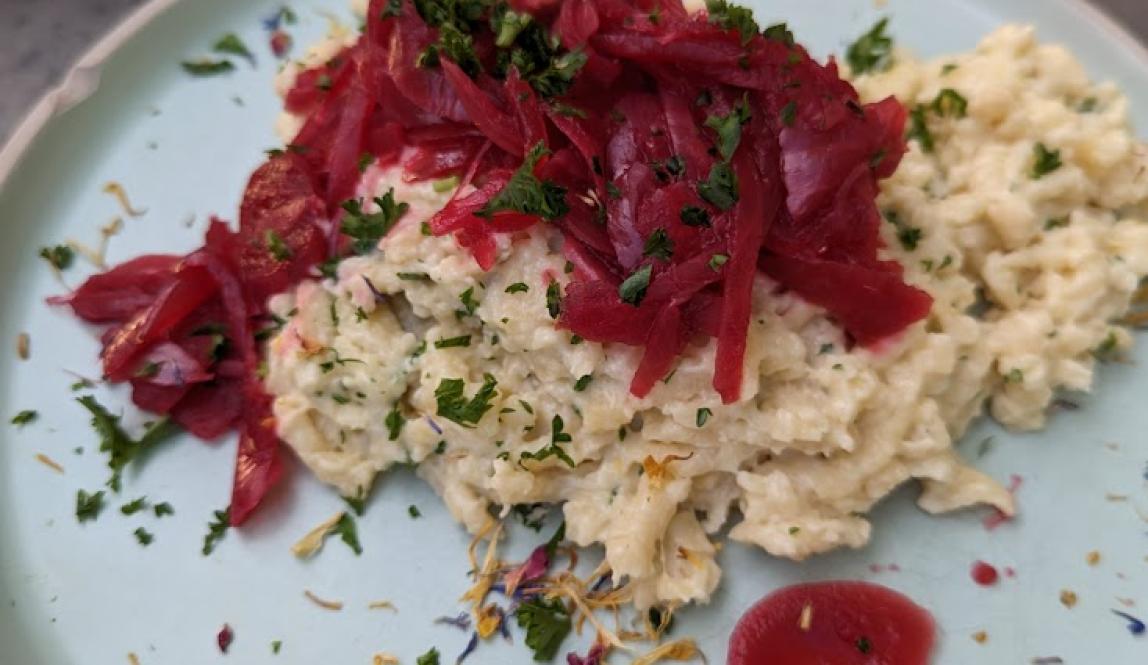 Pale yellow Spaetzle, resembling mac and cheese, sits on a light blue ceramic plate, the Spaetzle has pickled red onions on top along with green onion slices on top