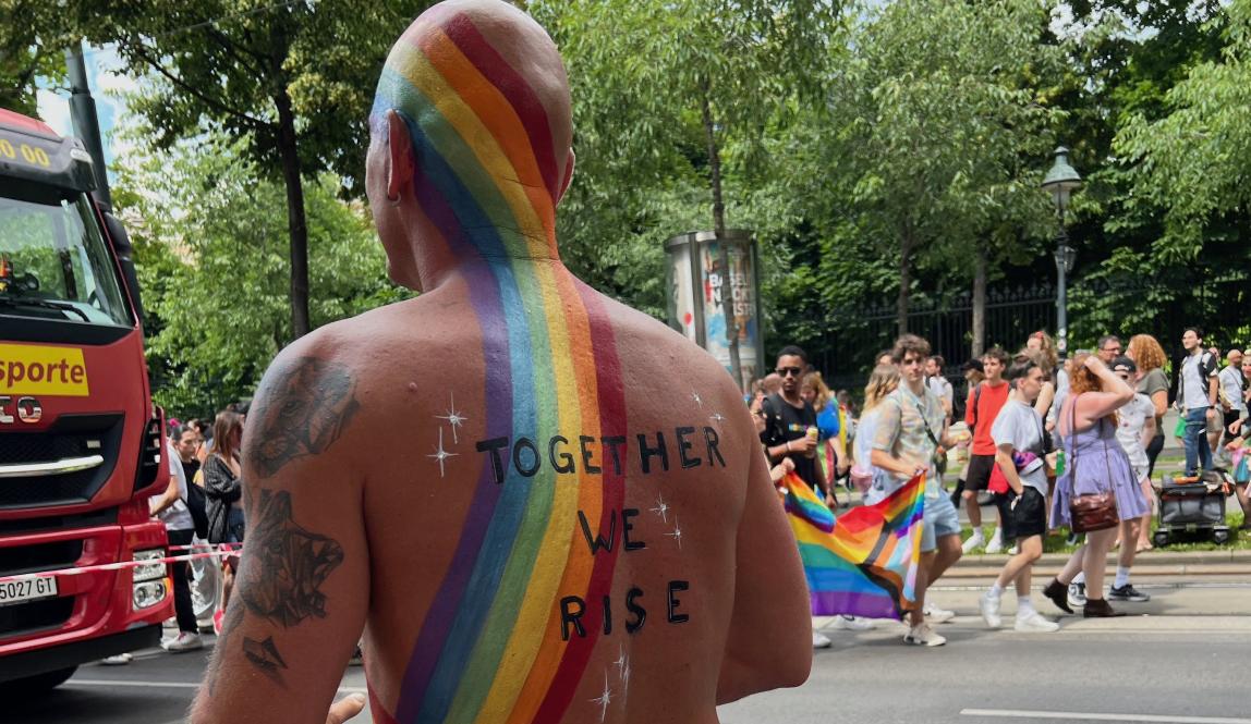 This is an image of a person attending the Pride Parade, with a rainbow painted across his back and the words "together we rise".