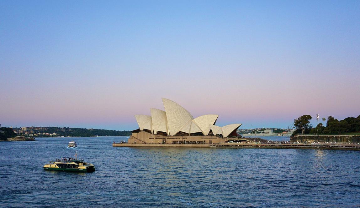 An aerial view of a boat sailing across Sydney Harbour in front of the Opera House at dusk