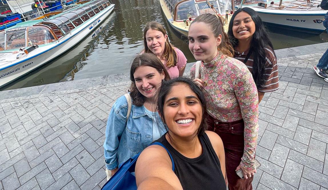five students standing in a line on cobblestone in front of a boat-filled amsterdam canal 