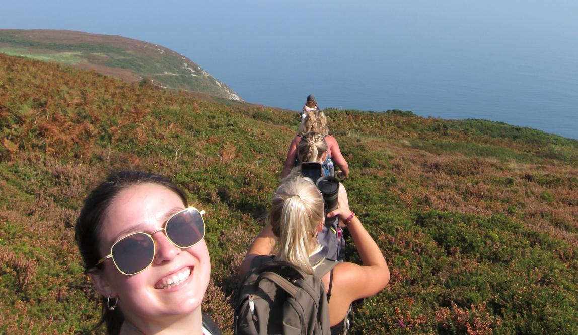 A line of students are hiking on a cliff, with the ocean in the background. The girl in front grins into the camera.