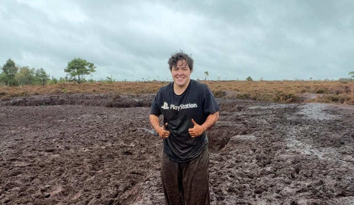 Short white man, dressed in dark muddy clothes, standing by a bog. He is smiling with two thumbs up