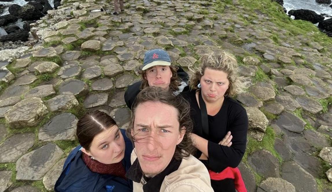 Four students taking a selfie on the rocks at Giant's Causeway, with the ocean visible in the background. They all are making silly faces. 