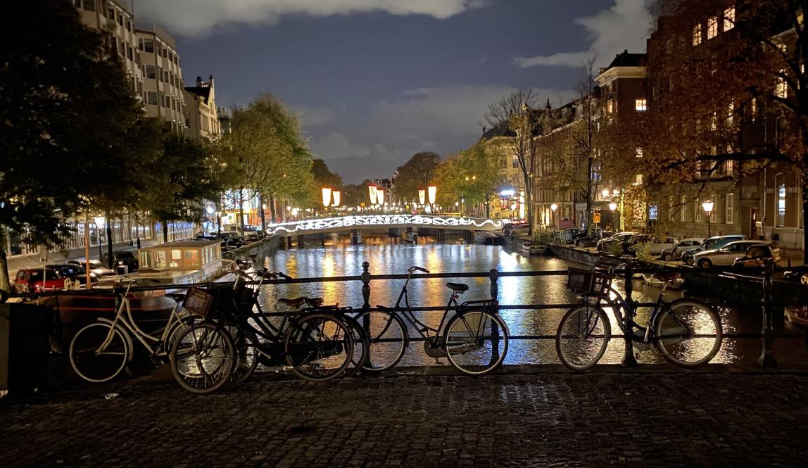 Bikes sitting by the rail of a canal in Amsterdam at night. The edges of a bridge in the distance are lit up, lighting the water and the scenery around.