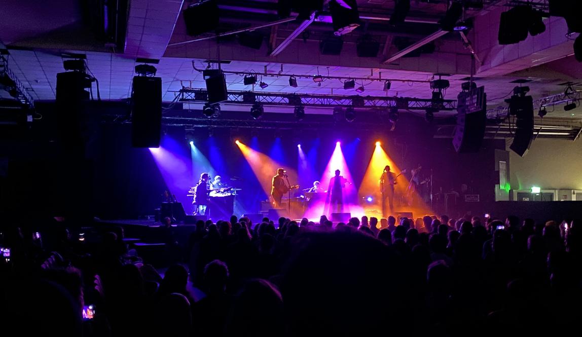 Gregory Alan Isakov, second from left, alongside his bandmates onstage, each in a different-colored spotlight. From the left is piano (blue), Gregory on guitar (orange), violin (pink), and bass guitar (yellow/orange). There is a sea of heads in front of them, an audience.