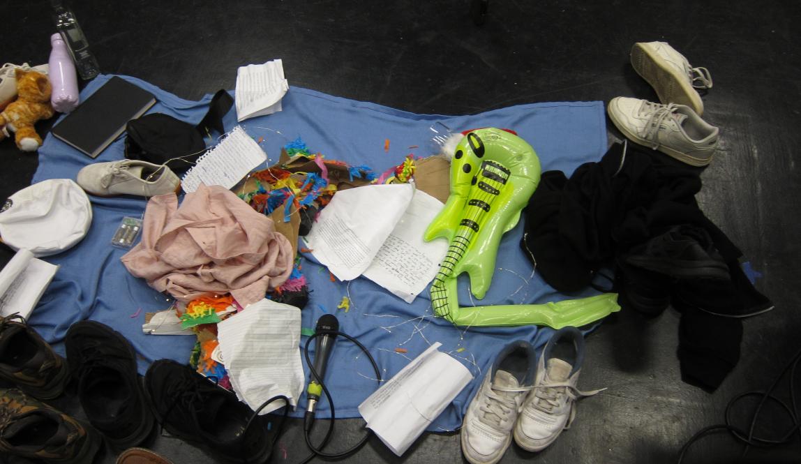 A messy pile of objects including shoes, a microphone, and a torn up piñata sit on a blue blanket on the floor.