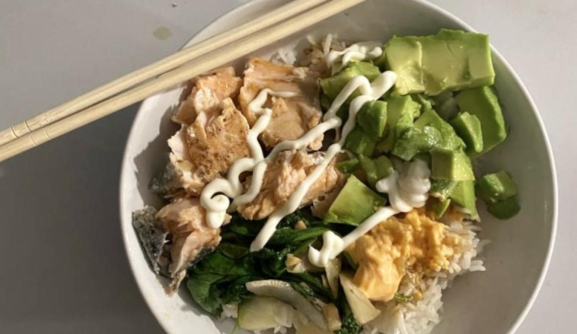 A bowl with chopsticks leaning on it, filled with fish, avocado, rice, and sauces. 