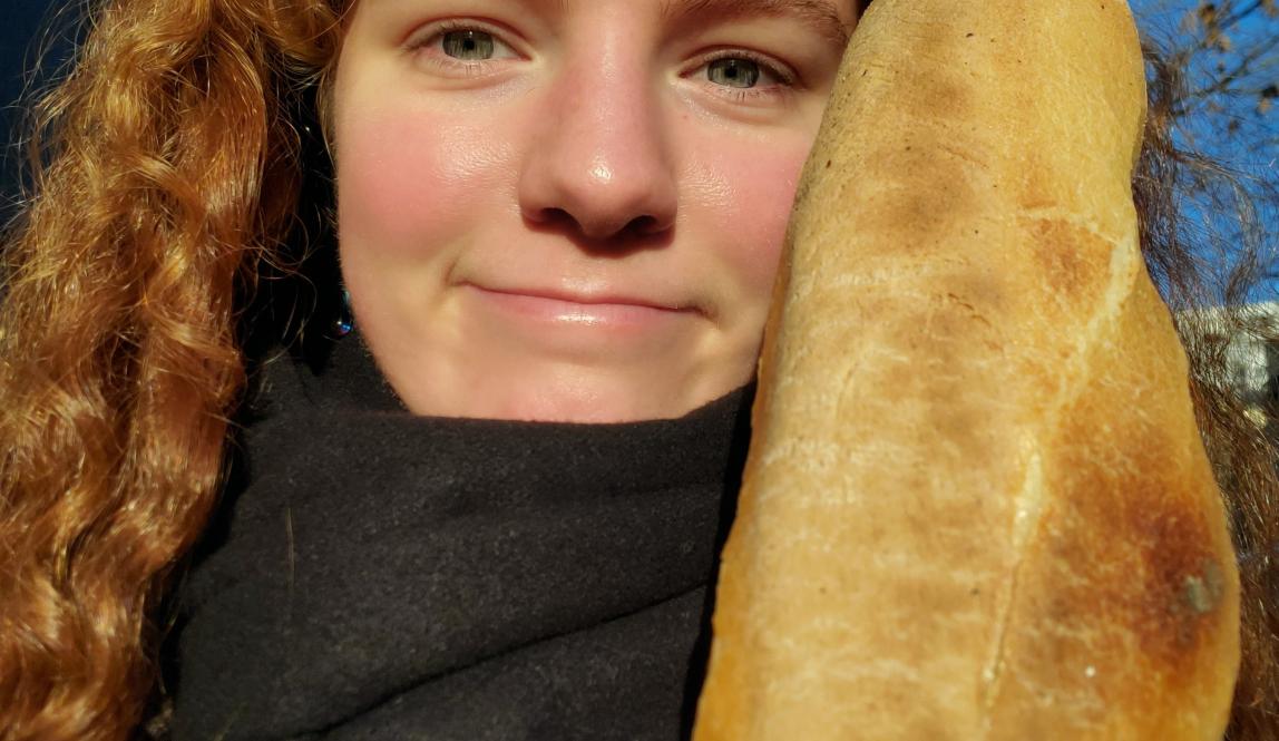 Ginger woman walking down the street smiling at the camera while holding a baguette in one hand.