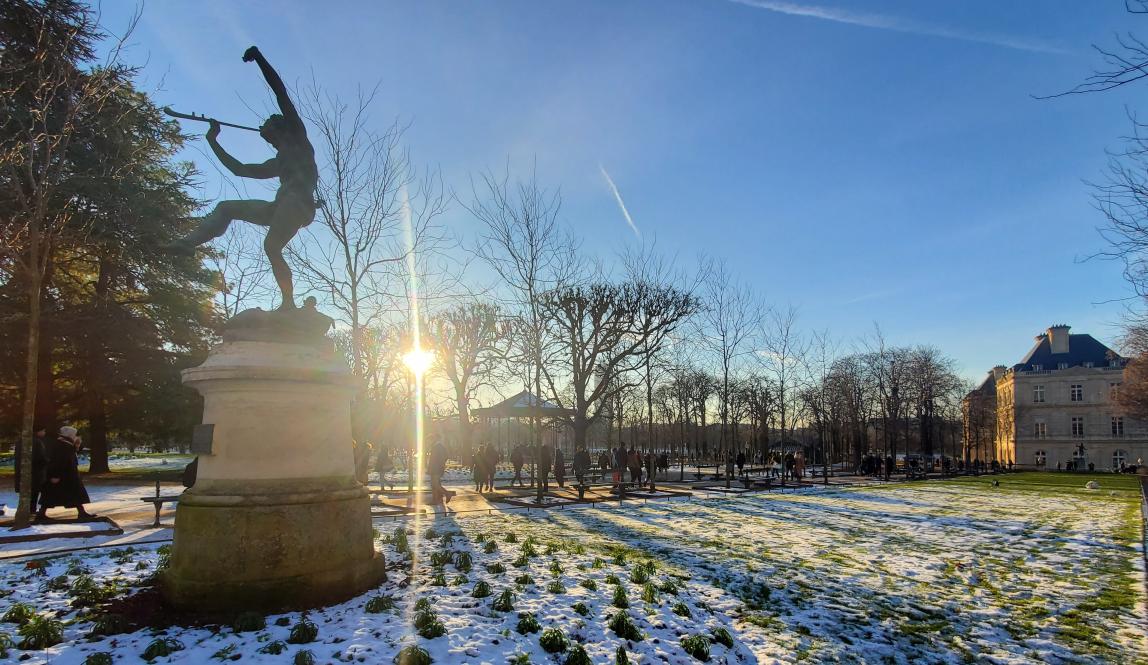 A statue in the Luxembourg Gardens with the sun behind it and green grass peeking out from under the snow.