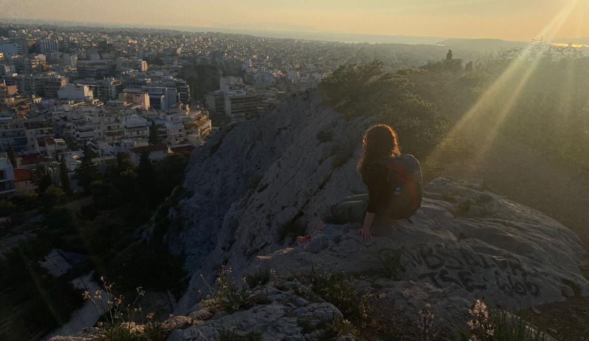 A ginger girl sitting on a cliffside, the sun setting behind her, while she contemplates seeing if she could fly