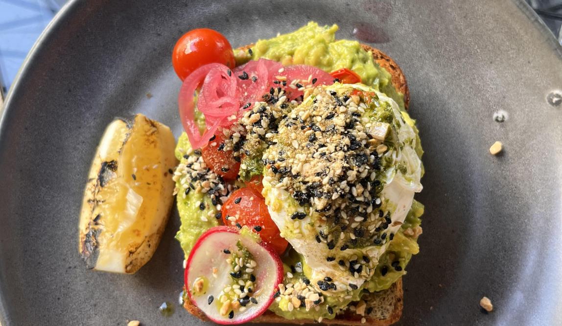 In this photo, there is a slice of cereal toast topped with avocado, tomato, lemon, and a poached egg from The Store in Auckland, NZ.