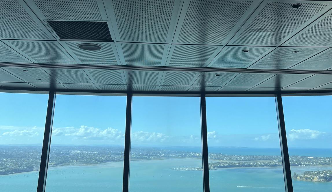 This photo shows the view from the Orbit 360 Dining Floor at the Sky Tower, featuring clear water outside and a dining table set with plates, napkins, and utensils inside.