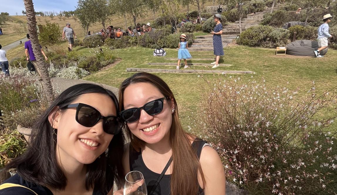 In this photo, two people pose for a selfie in an outdoor space at a vineyard. They both wear black sunglasses, black shirts, patterned skirts, and hold wine glasses in their hands. In the background, there are wooden steps, trees, shrubs, benches, bean bags, and people. The sky has a few clouds.