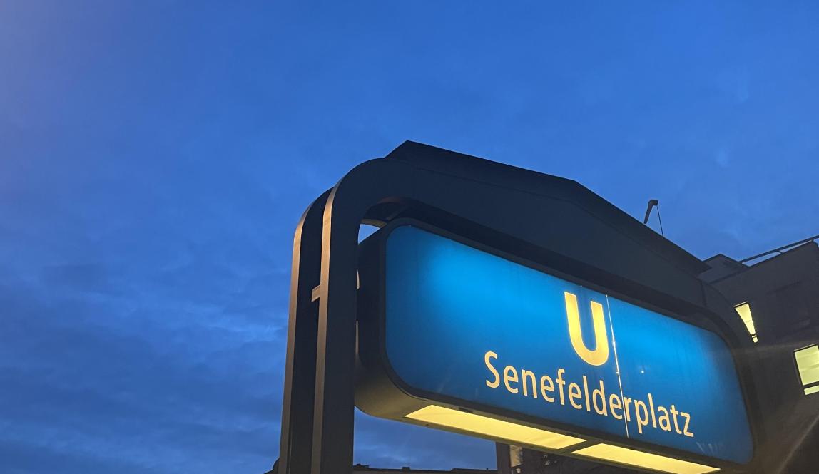 A blue U-Bahn station sign reading "Senefelderplatz" with a sunset in the background