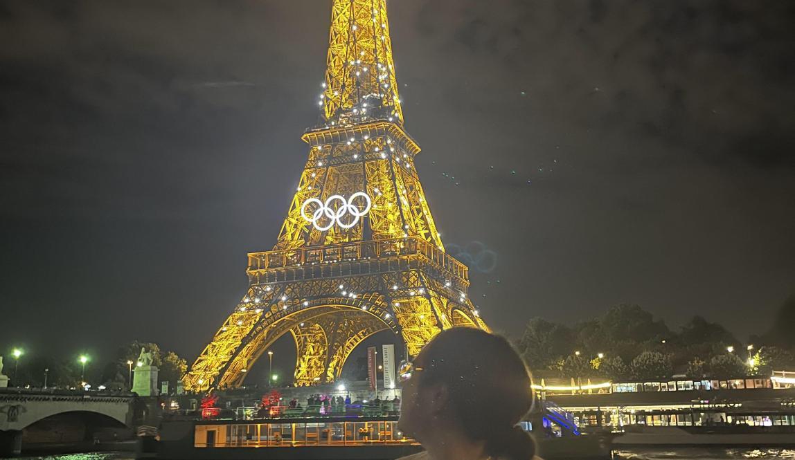 it is dark outside; the Eiffel Tower is in view across the Seine river and is sparkling; a girl stands and is admiring the tower with her head turned