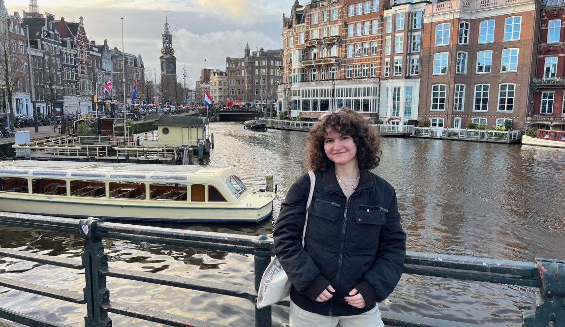 Student posing on a bridge above an Amsterdam canal, with a tour boat, waterfront buildings, and city landmarks visible behind them.