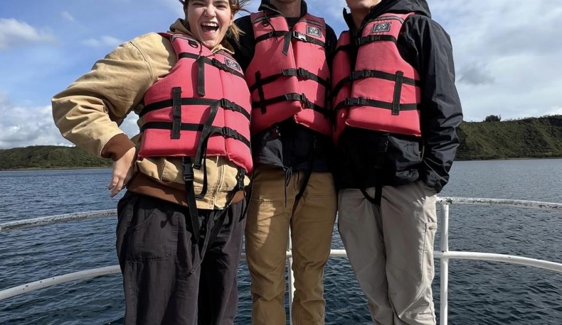 Three friends posed at the helm of a boat, standing. Lifejackets on and smiling.