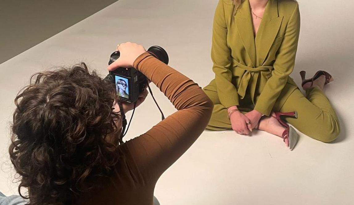 A fashion photoshoot with a model in a green pantsuit sitting on the floor, with the photographer sitting in front of them taking their photo