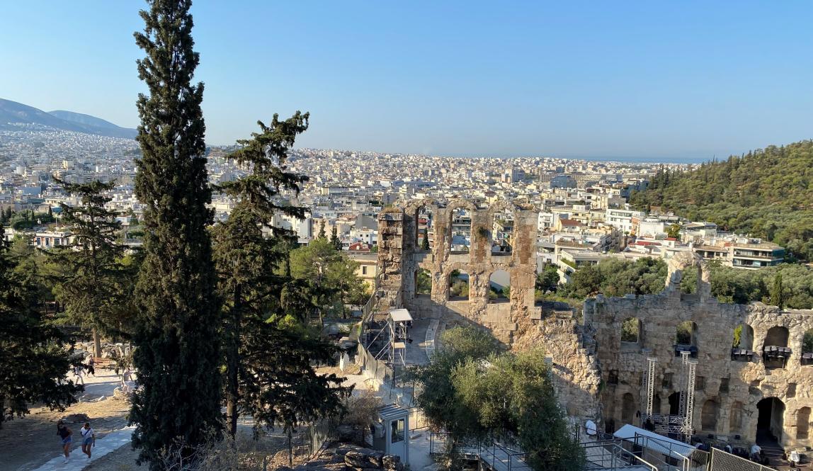 View of Acropolis ruins with cypress trees and cityscape in the background. This alt text was added with Al; accuracy may vary.
