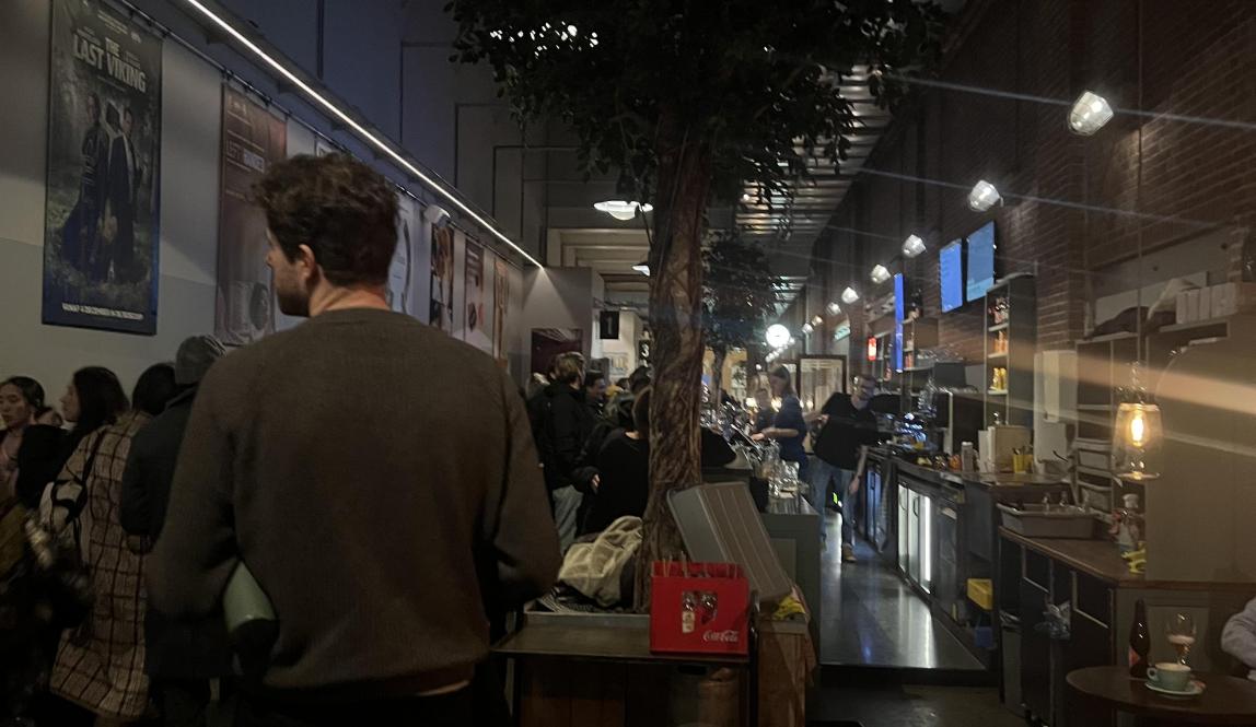 A crowded and busy hallway bustling with people as they order at the bar and sit at various tables.