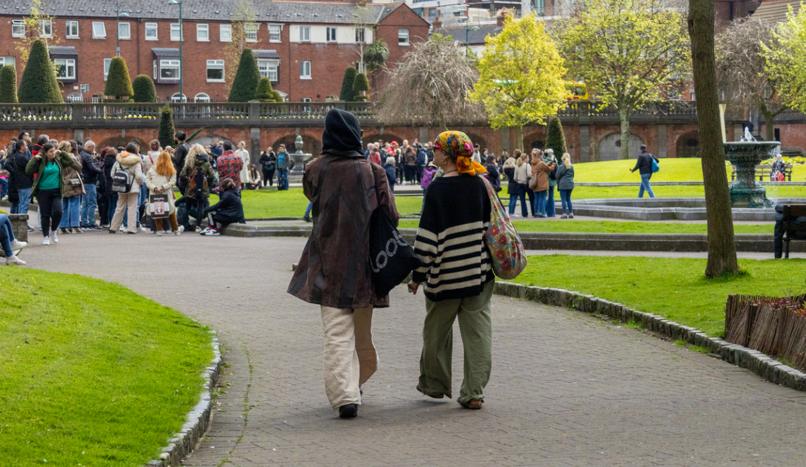 Two people walking through St. Patrick’s Park in Dublin, with a crowd gathered near a fountain and red-brick buildings in the background.