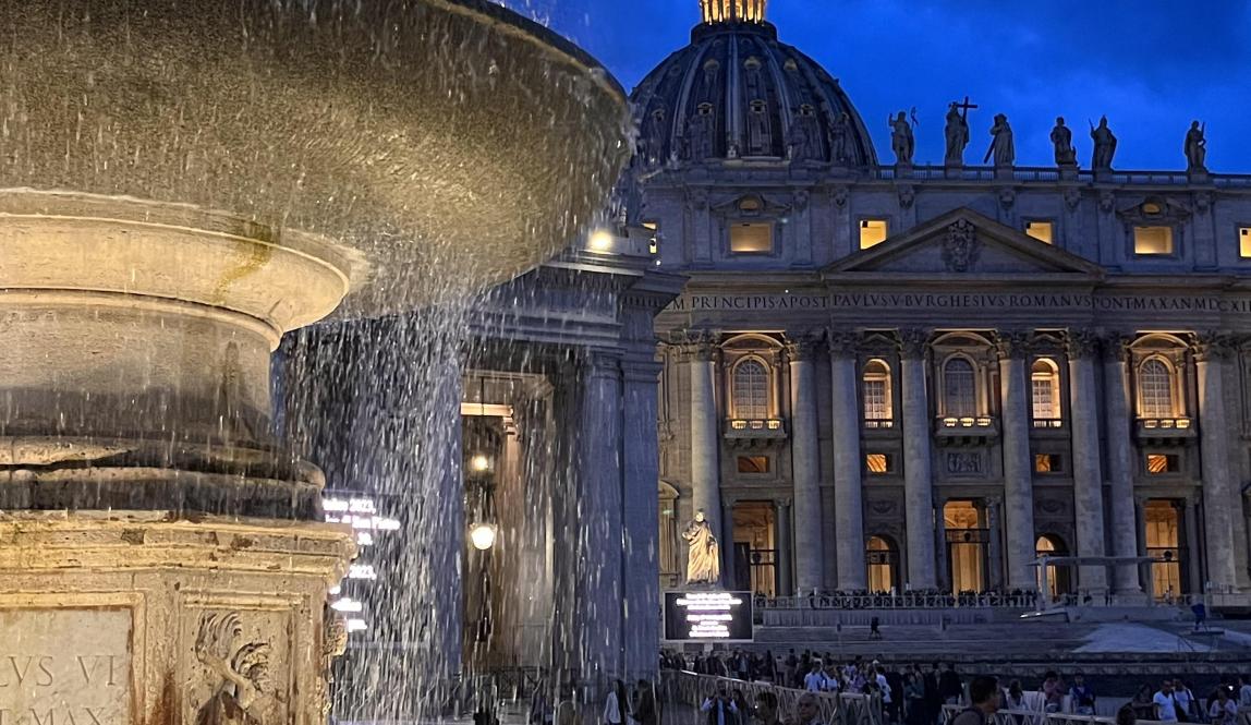 A nighttime photo of a fountain lit up in front of the Vatican.