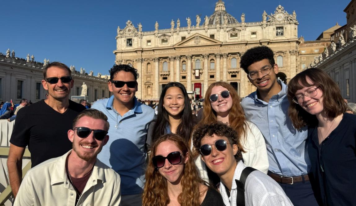 A group students wearing sunglasses and posing together on a bright, sunny day with historic architecture in the background
