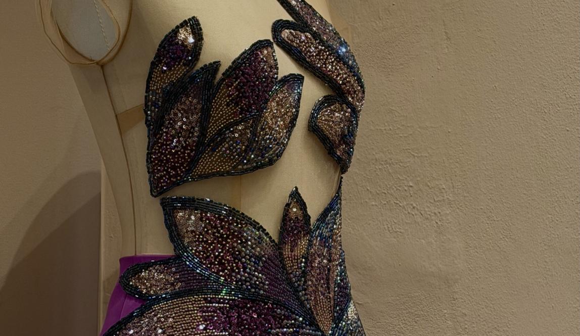 A photo of a gown with a sheer nude bodice and a purple skirt joined together with beaded appliqués that cover the bust and the waist. The beads are in gradients of gold, purple, and pink.   
