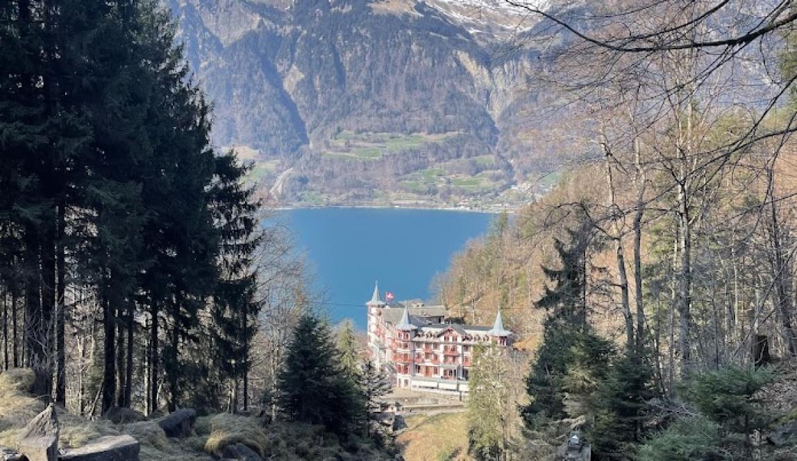 View of the lake in Brienz, an old hotel, and a massive mountain against a blue sky. Picture taken from a high elevation.