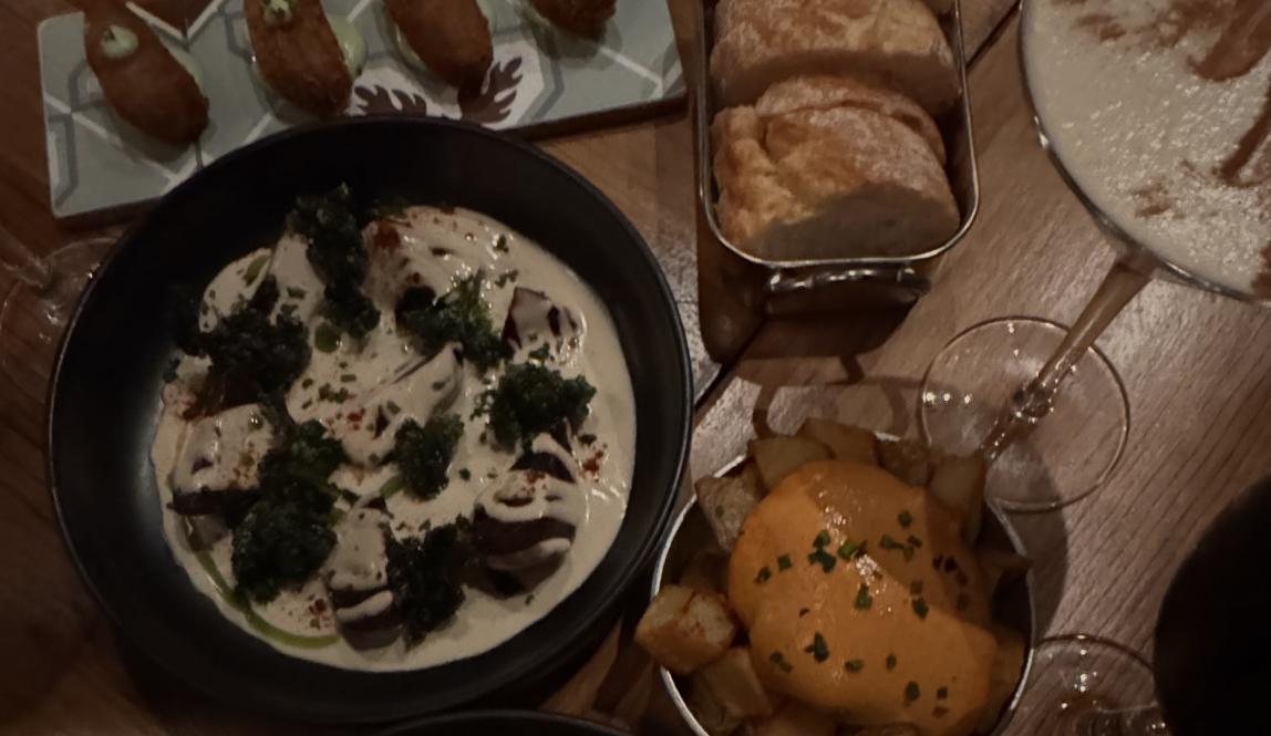 4 vegan croquettes next to a mini aluminum tray filled with bread. Below the croquettes is a dish composed of roasted beetroot in a cashew cream sauce along with patatas bravas in a tin pan. 