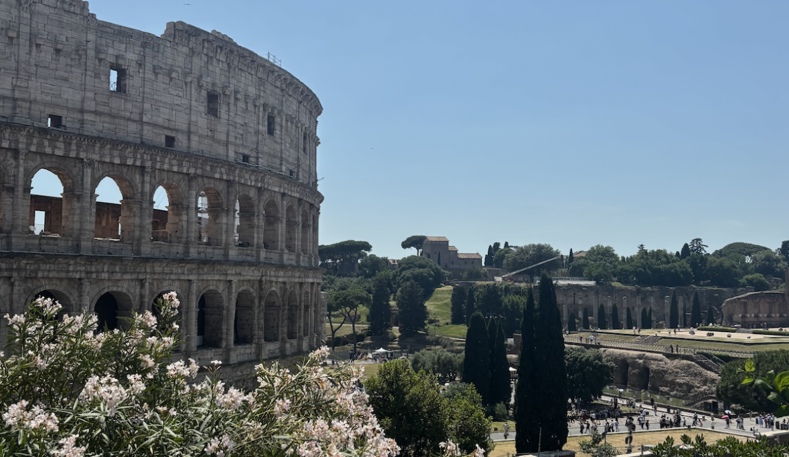 The famous Colosseum in Rome with its surrounding landscape of flowers, hills, trees, and tourists.