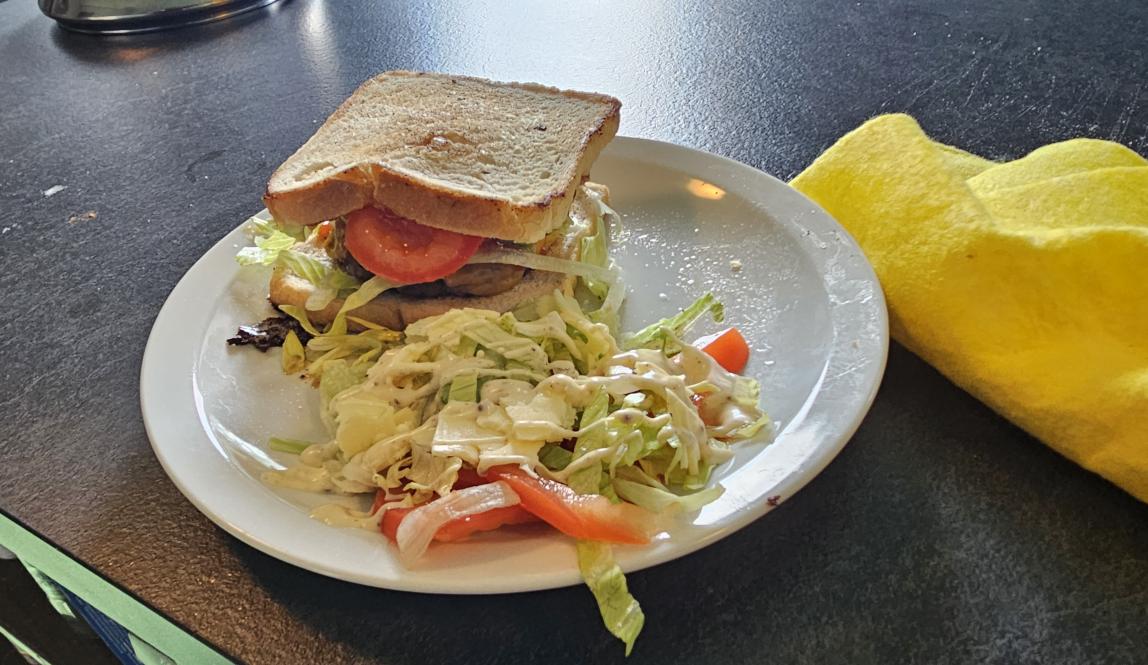 A sizeable beef burger with melted cheddar and young + mild cheese topped with sliced tomato, iceberg lettuce, sauced with barbeque. Accompanied with leftover lettuce and tomato for a side salad.