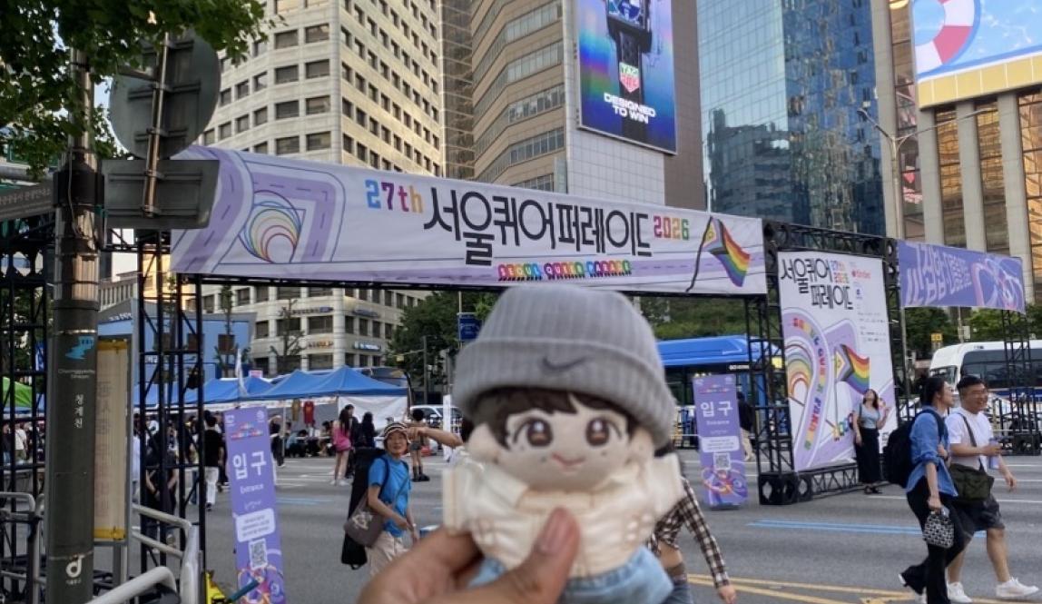 A hand holding up a plush doll in front of the entrance to the Seoul Queer Culture Festival.