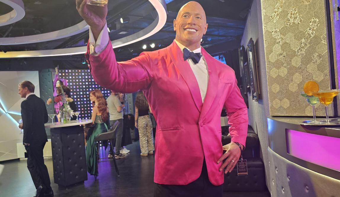 wax statue of the rock wearing a pink suit and black bowtie. he is also bald.