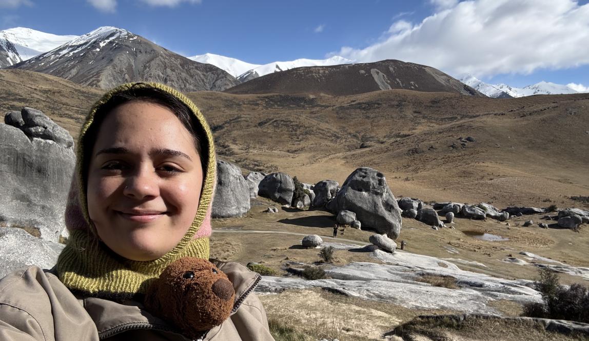 A photo of Pono in a windbreaker and balaclava and Teddy bear with the rocky dry landscape of Kura Tāwhiti behind.