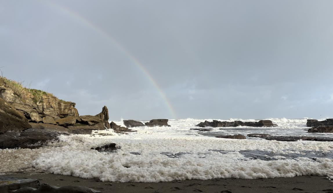 A rainbow over a seafoam covered black sand coast.
